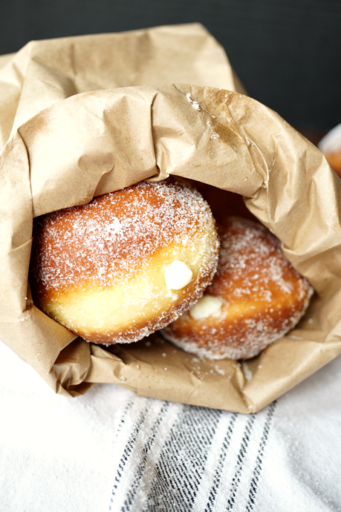 2 bomboloni, sugar-covered donuts filled with cream, inside a paper bag
