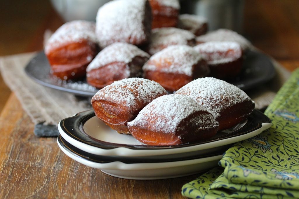Close up on 3 beignets, fried dark golden brown and covered with powered sugar, on a stack of plates. A big platter of beignets is in the background