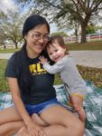 Katie seated on picnic blanket, baby Kyla standing holding onto her, both smiling